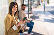 © Dorde - Smiling young woman looking away holding mobile phone while sitting at bus station and waiting for bus, using smart phone to check bus schedules, connections and maps, buying bus ticket on online app.