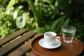 Wall Mural - White coffee cup and glass of water on wooden table with green lush background