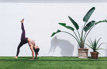 Wall Mural - Sportive fit girl in tracksuit stretching body for training muscles during terrace workout on weekend, active female athlete doing exercise for practicing own flexibility near decorative plant