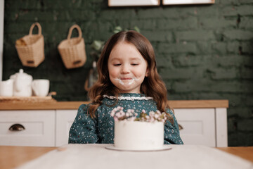 Adorable little child girl wears festive dress takes a bite out of a decorated flowers name cake at a birthday party. Happy smiling kid licks white cream from her dirty face and shows thumb up indoors