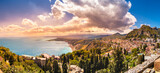 Fototapeta  - Taormina, Sicily, Italy. Panoramic view over Taormina town on hilltop, coastline of scenic bay in Ionian sea, Naxos town and mountains unter sunset sky. Popular tourist destination
