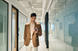 Young happy Asian business man using mobile cell phone tech standing in office hallway. Professional Japanese businessman holding smartphone, working on cellphone corporate device walking in office.