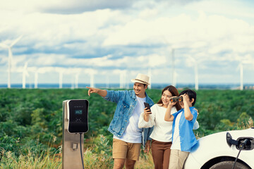 Concept of progressive happy family enjoying their time at wind farm with electric vehicle. Electric vehicle driven by clean renewable energy from wind turbine generator for charging station.