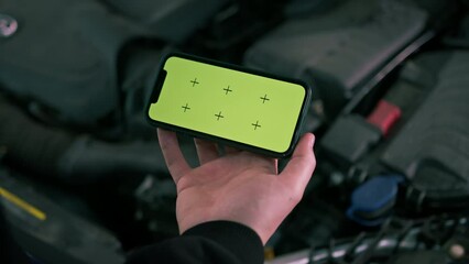 Male car service worker with a phone in his hands on which there is a green screen near the open hood of a luxury blue car