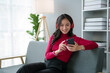 © amnaj - Cute Asian young woman wearing headphones sit on sofa and using smartphone. Asian woman red shirt is using mobile phone, watching movie, listening to music, shopping online, watching live broadcast.