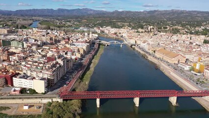 Wall Mural - Picturesque aerial view of Spanish city of Tortosa on banks of Ebro river with old red railway bridge and State bridge on sunny spring day, Catalonia. High quality 4k footage