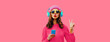 Trendy colorful portrait of stylish young woman in headphones listening to music with phone and blowing her lips sends air kiss on pink background