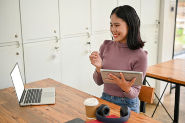 Wall Mural - A female college student remote working in the coffee shop, sitting at a table with her laptop, tablet