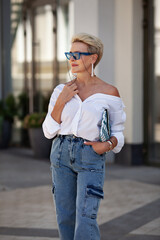 Beautiful mature woman with chic blonde bob outdoors in city street wearing trendy white shirt, cargo jeans pants and fashionable accessories, stylish glasses and clutch. Fashion style portrait