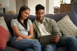 © StratfordProductions - Happy young biracial couple in casual clothing sitting on sofa using digital tablet at home