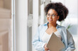 © JenkoAtaman - African american business woman standing with laptop near window in modern office