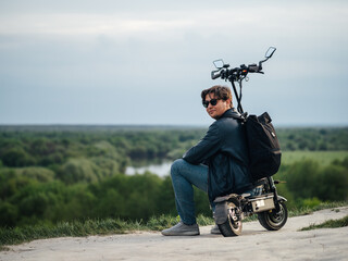 Wall Mural - A young man in sunglasses sits on a powerful electric scooter on a hill and admires nature. Summer lifestyle, modern electric transport