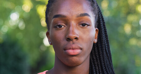 Wall Mural - Close Up of Beautiful Black Young Woman Looking at Camera with Neutral Expression with a Greenery Background. Happy Female Teenager Enjoying Fresh Air in the Park and Taking Care of her Wellbeing