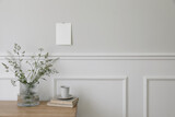 Fototapeta  - Elegant Scandinavian living room interior. Cup of coffee, books on wooden desk, table. Vase with green grasses and cow parsley. Blank greeting card, invitation mockup taped on wall. White moulding.