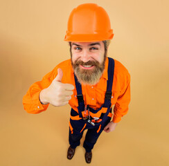 Wall Mural - Smiling bearded man in overalls and hardhat showing thumb up. Builder in safety helmet and uniform in full height. Male worker, repairman or builder in workwear with tools for repair. Advertising.