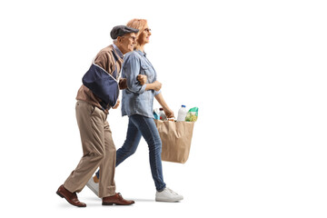 Wall Mural - Full length profile shot of a woman helping an elderly man with an injured arm and carrying grocery bags