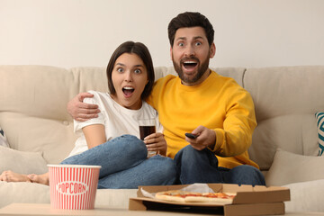 Canvas Print - Surprised couple watching TV with popcorn and pizza on sofa at home