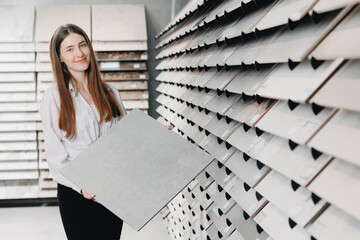 Seller Consultant young woman shows sample of gray ceramic tiles for loft style, refurbishment in an apartment bathroom