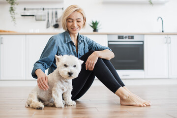 Wall Mural - Portrait of cheerful young lady sitting next to well-behaved white dog on bare floor in kitchen interior. Affectionate female keeper giving canine friend overall attention on bright sunny day.