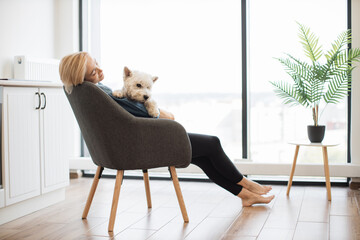 Wall Mural - Focus on West Highland White Terrier settled comfortably in pet lover's arms in light room with picture window. Smiling blonde female hugging small animal while stopping stressing out about job.