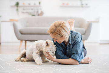 Wall Mural - Beautiful caucasian woman in cozy clothes resting on room floor together with white domestic animal. Charming young lady fondling entertaining pet's muzzle on carpet inside modern house.