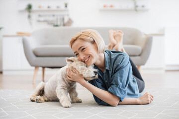 Wall Mural - Pretty smiling woman in casual wear snuggling to cute pet's head while testing comfort of wooden floor with carpet on it. Joyful healthy lady appreciating affection and support of her canine friend.