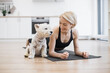 Healthy slim adult in sportswear holding arm balance yoga pose near white terrier sitting on black mat in kitchen. Caring pet lover communicating with canine buddy during Phalakasana II exercise.