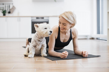 Wall Mural - Healthy slim adult in sportswear holding arm balance yoga pose near white terrier sitting on black mat in kitchen. Caring pet lover communicating with canine buddy during Phalakasana II exercise.