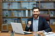 © Liubomir - Portrait of mature senior businessman in office, man looking at camera and holding hands up super power and superhero gesture, successful investor using laptop inside building.