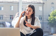 © puhimec - A young woman is talking on a smartphone and looking at a laptop screen.