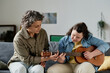 © AnnaStills - Teacher teaching girl with down syndrome to play guitar while they sitting on sofa in the room