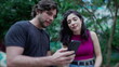 © Marco - Young man showing online content to woman on cellphone device, both standing outside at city park. Two people looking at smartphone screen comenting on the media they are watching