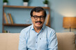 © WESTOCK - Happy indian senior man with eyeglasses by looking at camera while sitting on sofa at home - concept of successful, confident and satisfaction.