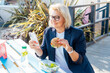 © okrasiuk - Middle-aged business woman eating a wholemeal burger with salad and using a phone sitting at picnic table in the park while having a break. Healthy lunch box for weight loss. Balanced diet.