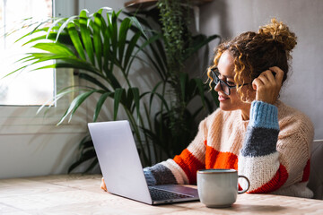 Adult woman at home surfing the net using laptop on the table with big plant in background. Home indoor technology leisure activity. Modern people lifestyle reading email on computer. Smart working
