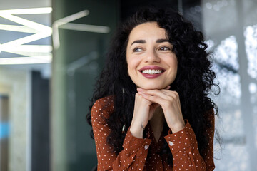 Beautiful and successful business woman inside office close up smiling and looking out the window, latin american boss dreams about successful achievement results in workplace.