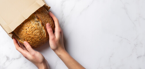 Fresh sourdough bread in woman's hands, top view. Woman takes a loaf of rustic wheat bread from a paper bag. Copy space banner on white marble background.