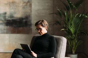 Middle-aged businesswoman in black formal wear sitting indoors holding a digital tablet