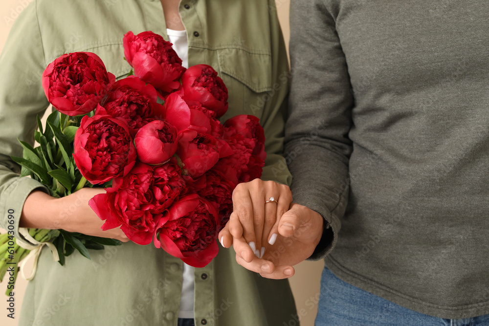 Happy engaged couple with flowers holding hands on beige background, closeup