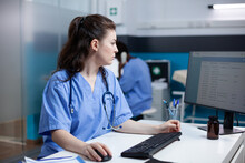 Caucasian Nurse Checking Appointments List On Computer Monitor, Sitting At Clinic Desk. Focused Adult Woman Healthcare Expert Working In Medical Office With Stethoscope Around Neck