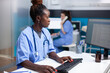 © DC Studio - African american nurse working at desk, inserting data in appointment table list. Concentrated adult woman checking patient list, wearing stethoscope in clean, modern medical office operating computer