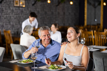 Wall Mural - Smiling senior man spending time with young girl in restaurant. Carefree couple laughing merrily, enjoying delicious food and drinking wine at dinner. Age-gap relationships concept