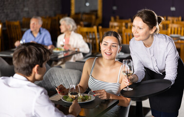 Wall Mural - Polite smiling waitress serving ordered drinks to young couple sitting at table, visiting restaurant for romantic dinner