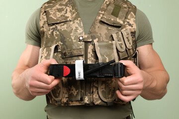 Wall Mural - Soldier in military uniform holding medical tourniquet on light olive background, closeup