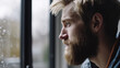 © wetzkaz - young adult man with light blond hair and a full beard, at home by the window, looking out of the window, rainy gray cloudy weather
