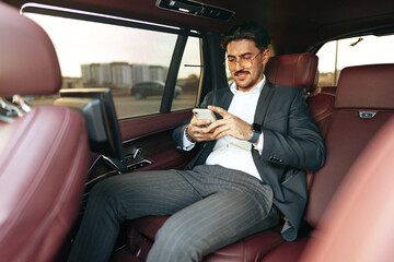 Young elegant businessman sitting on the back seat of a luxury car and using smartphone