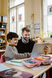 © Maskot - Happy male teacher assisting schoolboy using tablet PC at desk in school