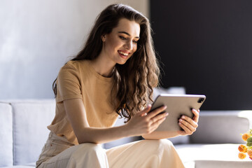 Happy young woman sitting on sofa and using tablet pc in apartment