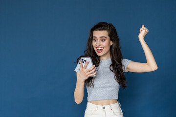 Image of a happy surprised young woman isolated over blue wall background using mobile phone make winner gesture.