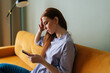 © dikushin - Close-up side view of sad young woman sitting on yellow couch looking at mobile phone screen, holding head, thinking about problem, feeling failure and exhausted, suffering grief and bad relationship